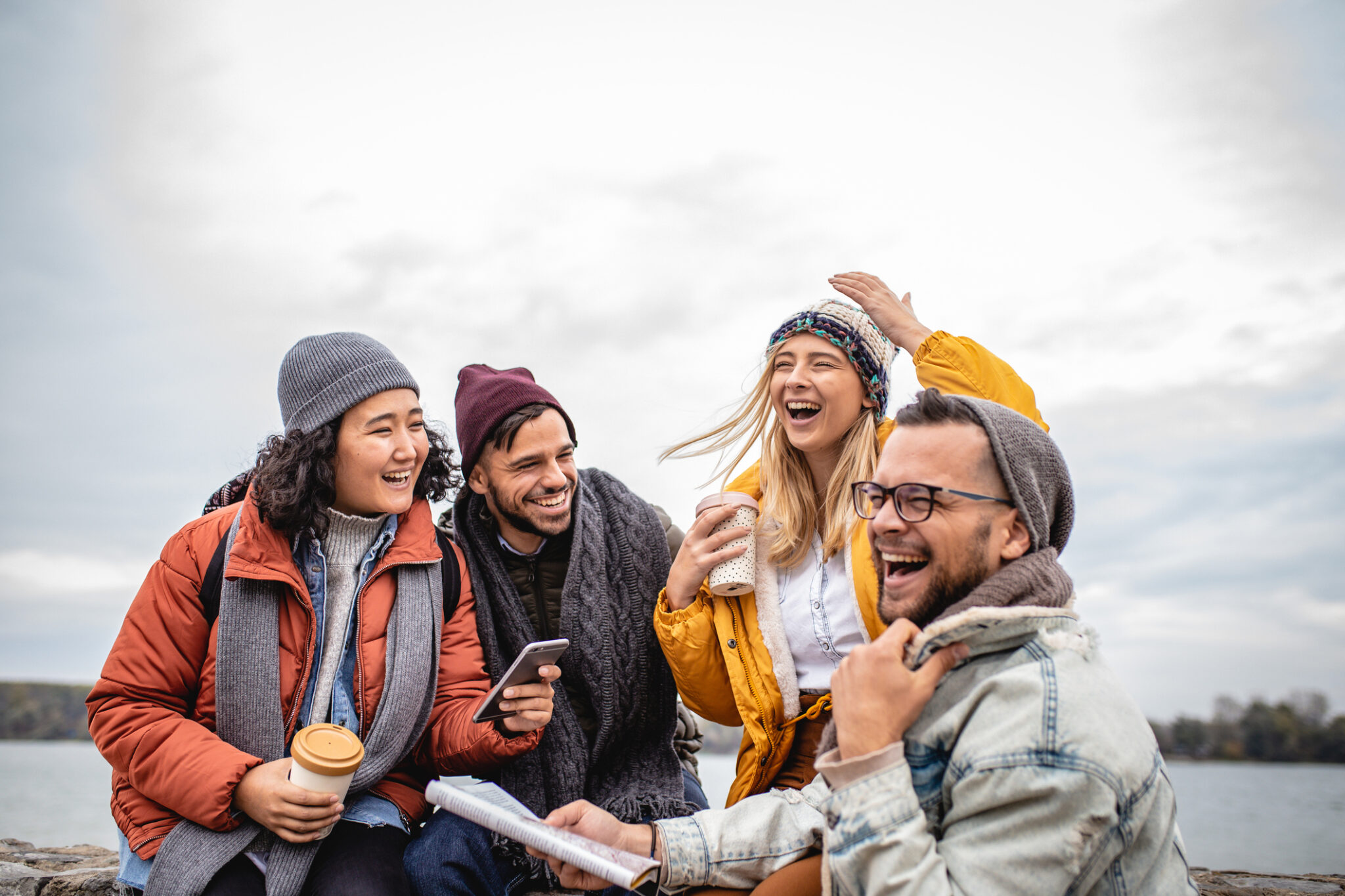 A group of four young friends is on the city break. They are laughing and sitting by the river